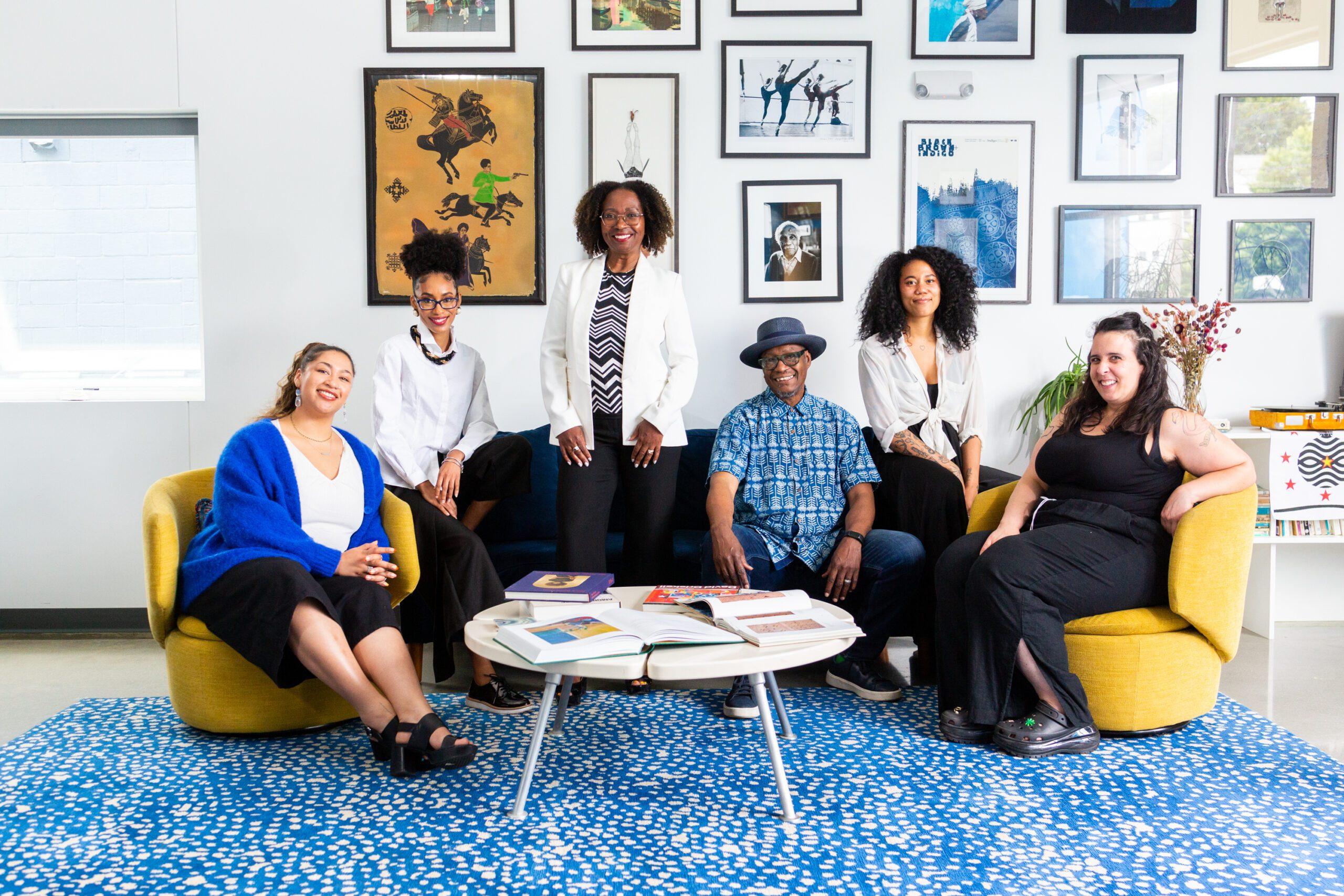 A group of people stand in a group smiling, surrounded by art on the walls and a colorful blue rug below.