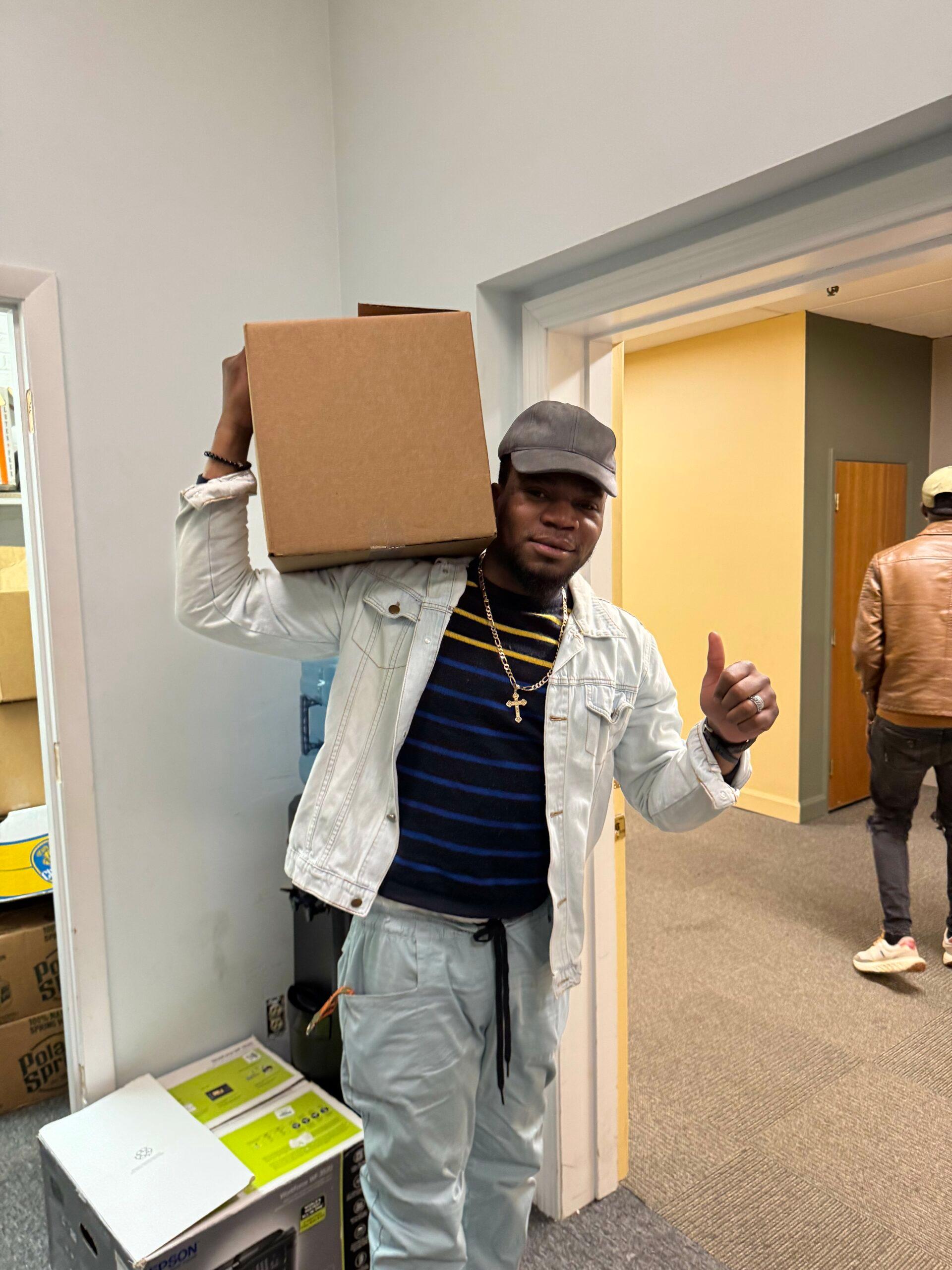 A person carrying a box on one shoulder while giving a thumbs up with the opposite hand