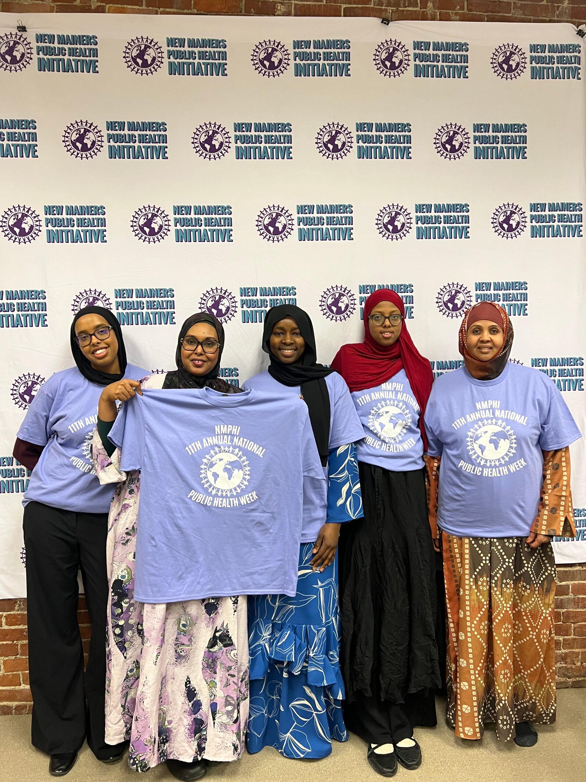 Five women stand together in front of a banner, wearing matching t-shirts