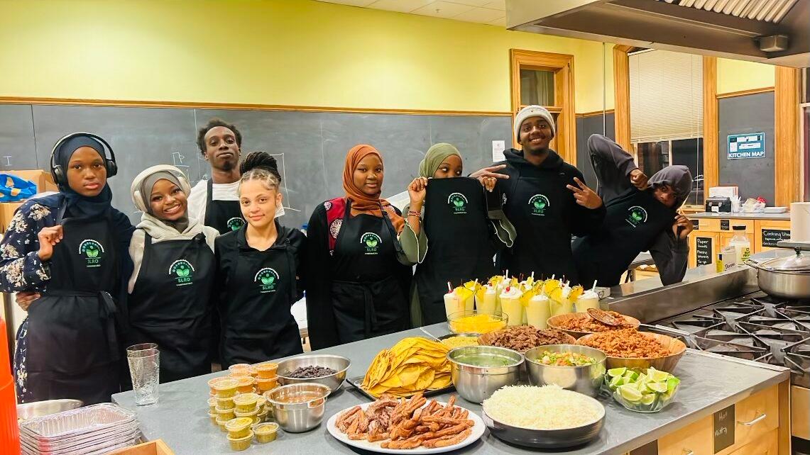 Young people wearing aprons stand alongside a table filled with food