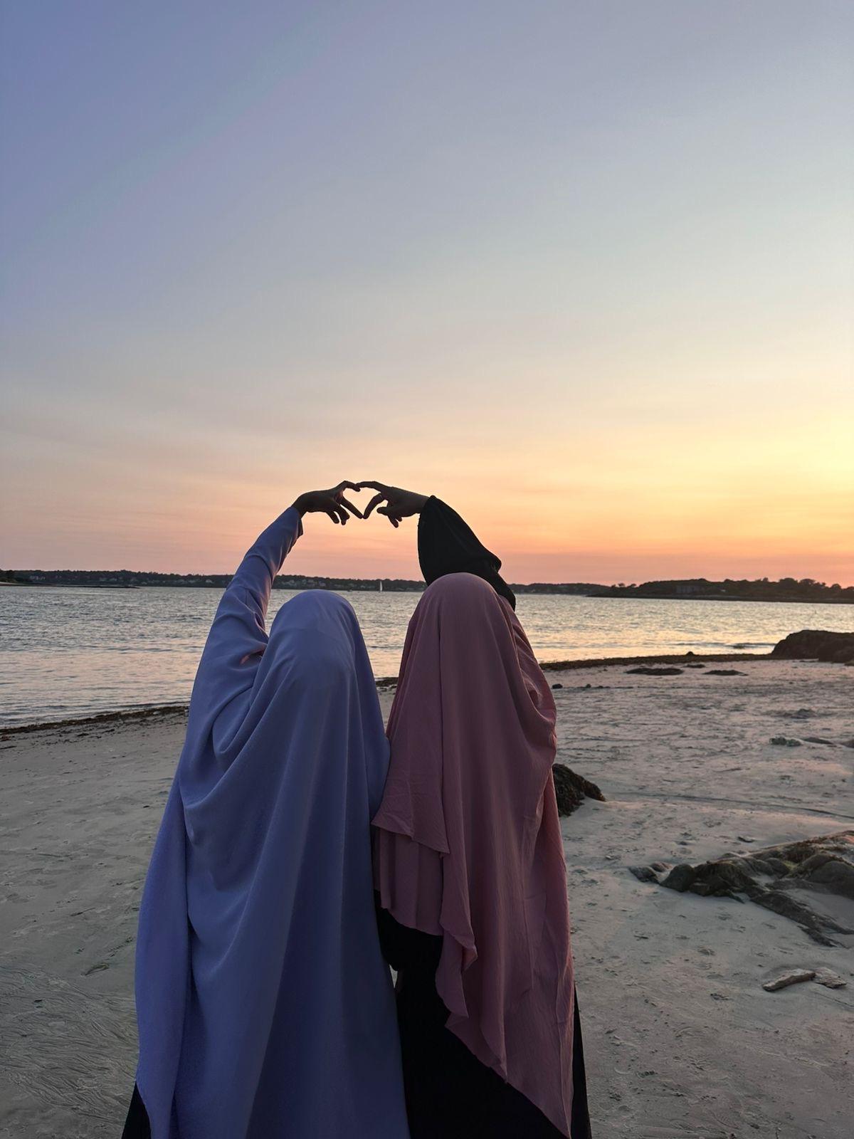 Two people stand next to each other on the beach at sunset, making a heart shape with their hands