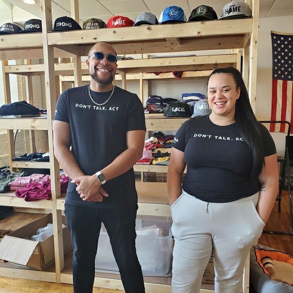 Two black adults stand and smile with black t-shirts saying, don't talk, act.