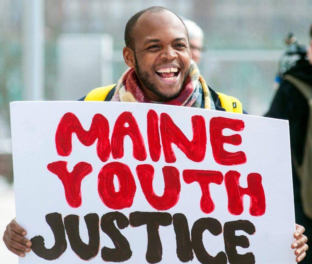 A smiling man holds a sign saying, Maine Youth Justice